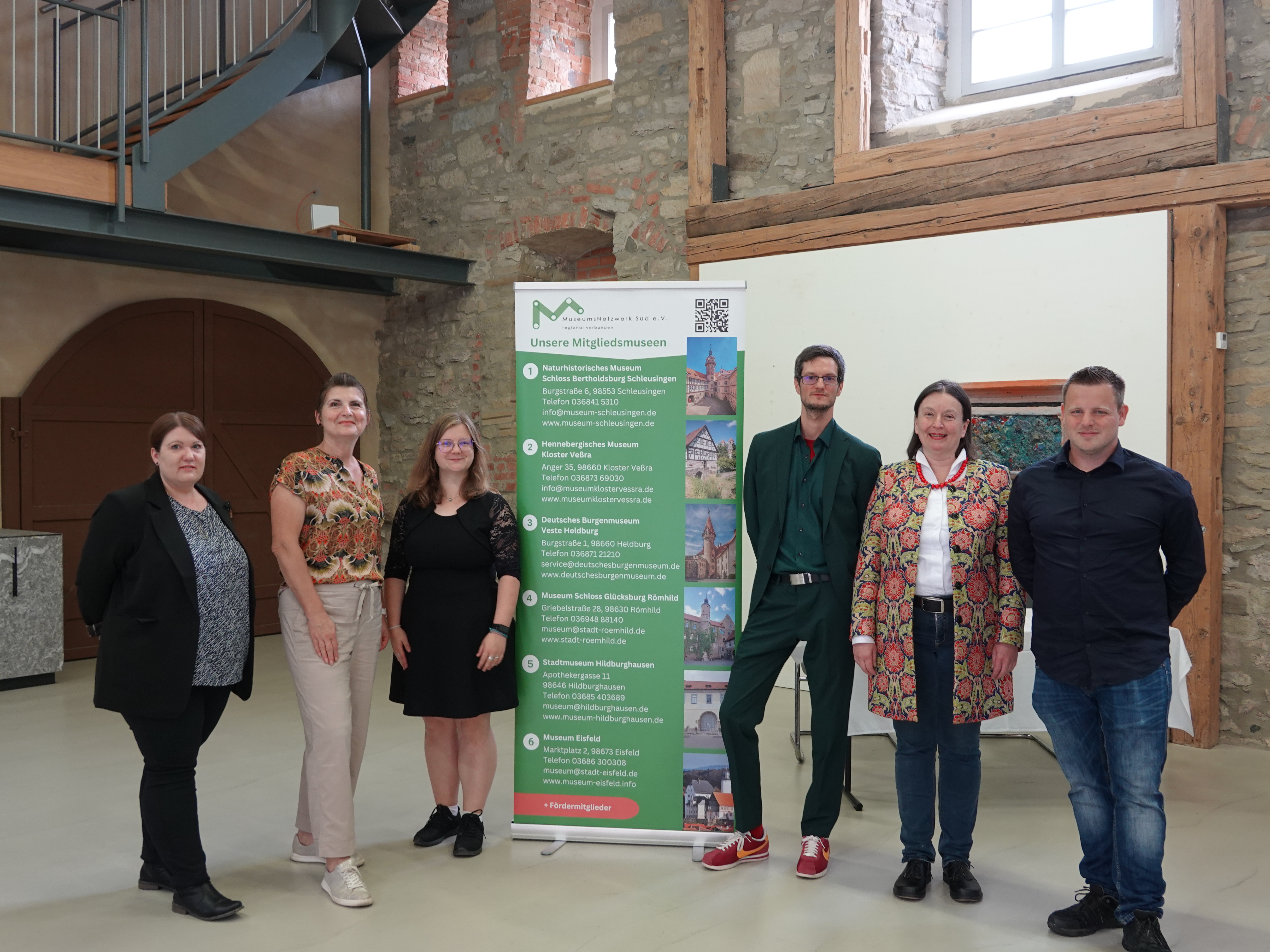 Gruppenfoto mit allen Mitarbeitenden des MuseumNetzwerk Süd e. V., links und rechts neben einem Banner mit den Mitgliedsmuseen stehend